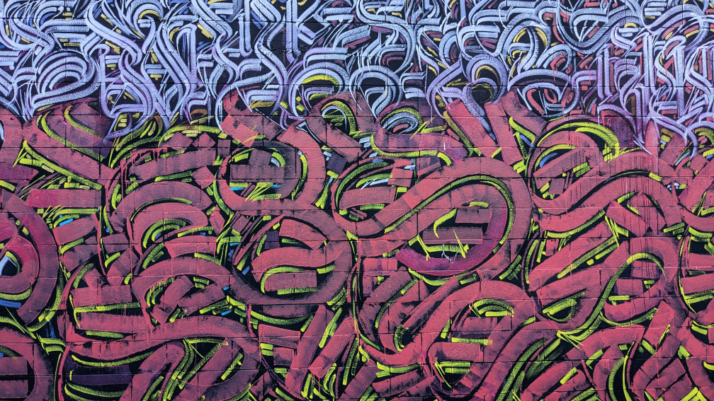 This is a close-up shot of a vibrant graffiti mural on a brick wall, divided into two distinct horizontal sections of abstract, calligraphic-style art; the lower two-thirds of the mural features thick, flowing, and overlapping lines in a deep raspberry red, outlined in black and highlighted with strokes of bright lime green, creating a sense of depth and energetic movement, while the upper third consists of a more dense and intricate pattern of thinner, interwoven lines in shades of purple, white, and black [gen AI description].